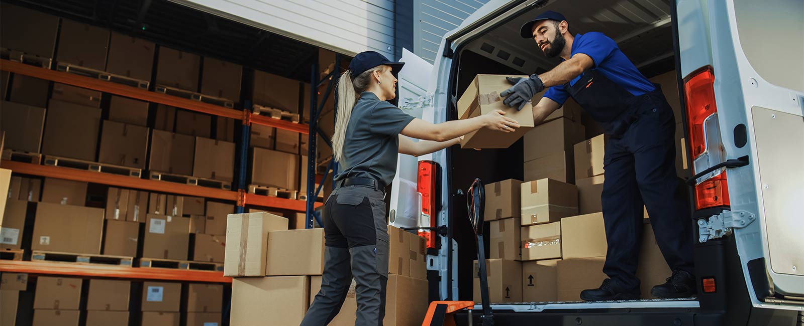 A man in a van full of boxes hands one box to a female warehouse worker.
